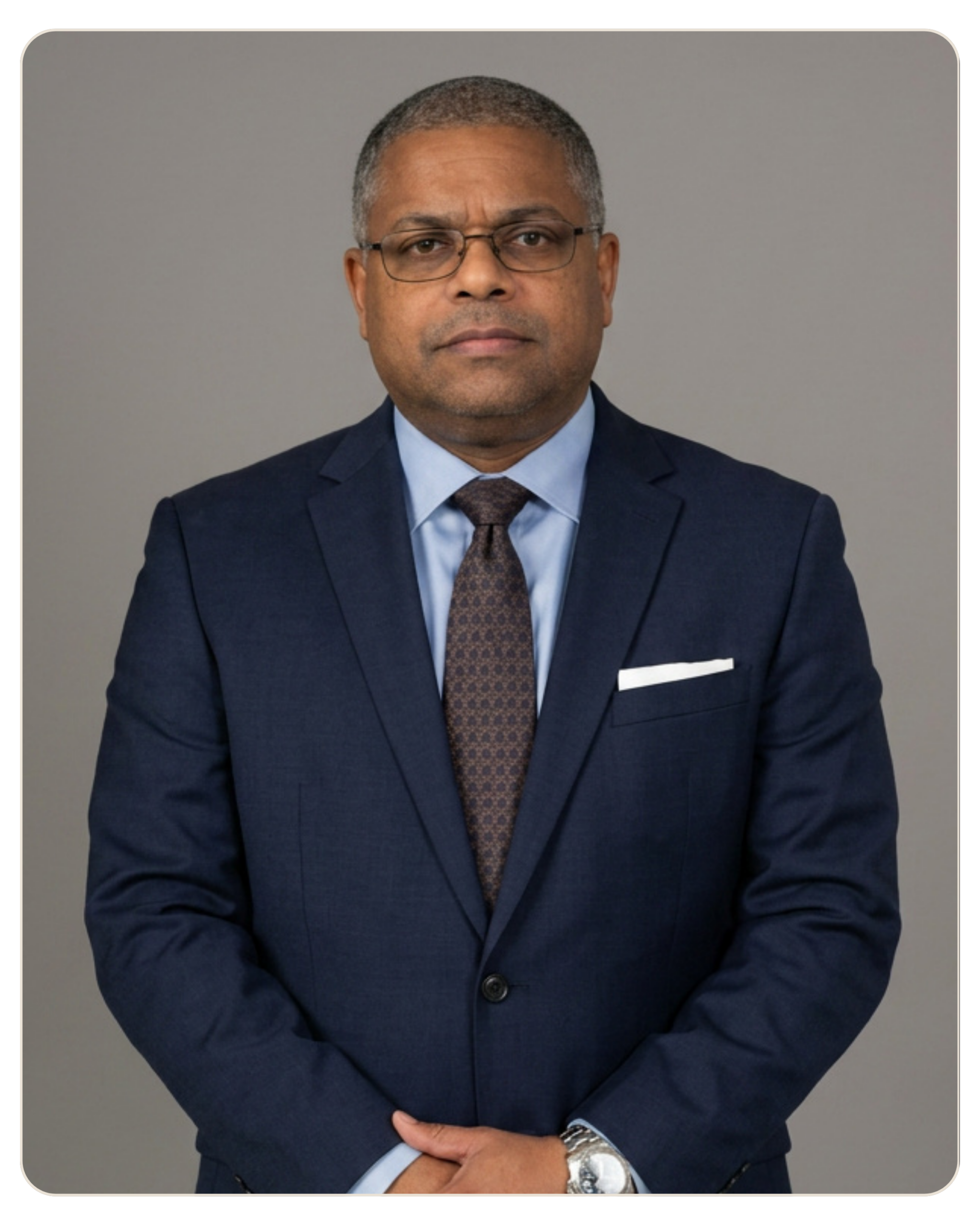 Flyland - Trost - Anthony Bundy, LPC Professional portrait of a middle-aged man in a navy suit, light blue shirt, patterned brown tie, wearing glasses against a gray background.